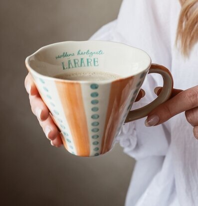 Bild på krämfärgad mugg med orange och blå ränder i handen