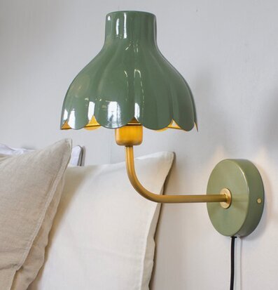Bild på stilren vägglampa i mossa och guld ovanför säng